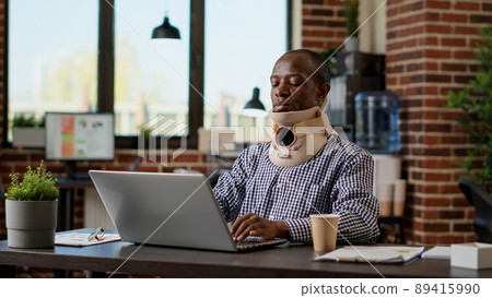 Financial analyst with physical injury wearing medical neck collar at office job, using cervical foam to treat vertebrae accident. Recovering from painful spine injury. Tripod shot. Financial analyst with physical injury wearing medical neck collar at office job, using cervical foam to treat vertebrae accident. Recovering from painful spine injury. Tripod shot. 89415990