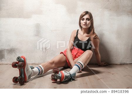 Attractive woman sitting on the floor in roller skating looking at camera. Studio shot, loft interior Attractive woman sitting on the floor in roller skating looking at camera. Studio shot, loft interior 89418396