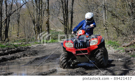 A girl driving a red quad bike rides through a puddle in the spring forest. Front view. Extreme type of outdoor activities. ATV riding. A girl driving a red quad bike rides through a puddle in the spring forest. Front view. Extreme type of outdoor activities. ATV riding. 89421437