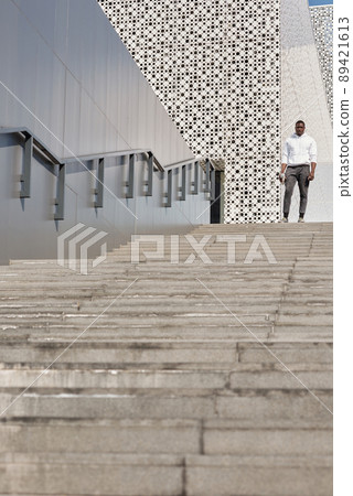 Athletic Black man in stylish sportswear standing on concrete stairs against contemporary office building with perforated facade, low angle view Athletic Black man in stylish sportswear standing on concrete stairs against contemporary office building with perforated facade, low angle view 89421613