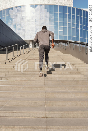 Unrecognizable athletic Black man running up concrete stairs towards modern business center while having outdoor workout, low angle rear view 89421665