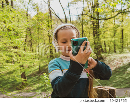 Adorable child girl in headphones listening to music using smartphone outdoor sunny day. Kid with gadget in the park. Adorable child girl in headphones listening to music using smartphone outdoor sunny day. Kid with gadget in the park. 89421765