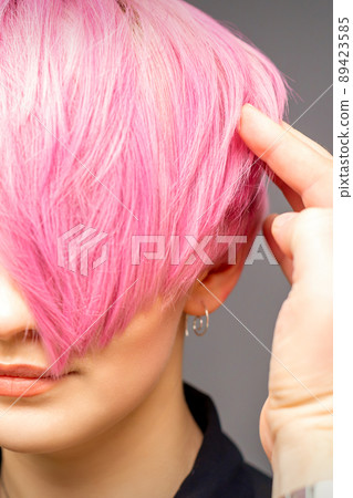 Hairdresser with hands and comb is checking out and fixing the short pink hairstyle of the young white woman in a hair salon 89423585
