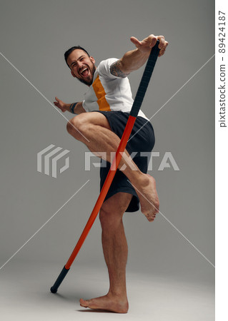 A fighter in white t-shirt and black shorts, stands in a fighting artificially pose and holds a fight stick on studio background. 89424187
