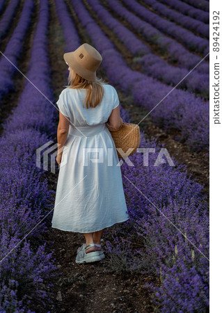 A middle-aged woman, blonde in a white dress and hat, walks through a lavender field with a basket 89424192