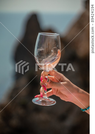 A female hand holds a glass of wine against the background of the sea and mountains. A female hand holds a glass of wine against the background of the sea and mountains. 89424194