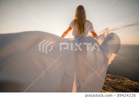 A beautiful young woman in a white light dress with long legs stands on the edge of a cliff above the sea waving a white long dress, against the background of the blue sky and the sea. A beautiful young woman in a white light dress with long legs stands on the edge of a cliff above the sea waving a white long dress, against the background of the blue sky and the sea. 89424216
