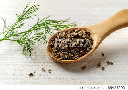 Whole dill seeds in a wooden spoon and fresh green dill sprig on a white wooden table. 89425503
