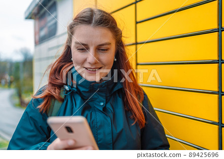 Caucasian woman showing smartphone with smart delivery app near a yellow automatic post terminal.  89425696