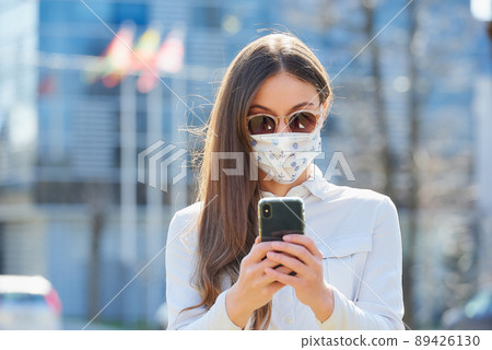 A woman wears a face mask to avoid the spread of coronavirus using a smartphone. 89426130