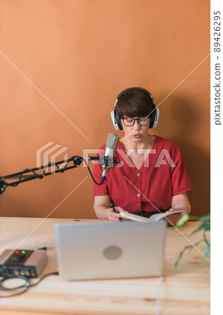 Middle-aged female radio presenter talking into the microphone and reading news - radio broadcast online concept Middle-aged female radio presenter talking into the microphone and reading news - radio broadcast online concept 89426295