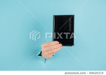 A woman's hand sticks out of a hole in a blue paper background and holds a passport in a black cover. A woman's hand sticks out of a hole in a blue paper background and holds a passport in a black cover. 89426314