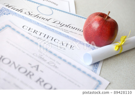 A honor roll recognition, certificate of achievement and high school diploma lies on table with small scroll and red apple. Education documents A honor roll recognition, certificate of achievement and high school diploma lies on table with small scroll and red apple. Education documents 89428110