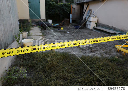 Victim of a violent crime in a backyard of residental house in evening. Dead man body under the yellow police line tape and evidence markers on crime scene 89428278