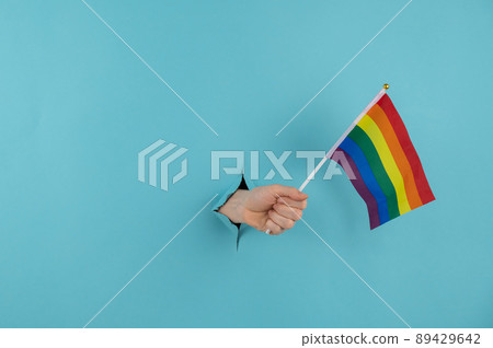 A woman's hand sticks out of a hole in a blue paper background and holds an lgbt flag. A woman's hand sticks out of a hole in a blue paper background and holds an lgbt flag. 89429642