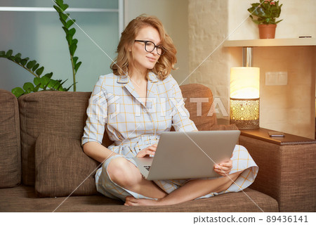 A young woman in glasses works remotely on a laptop in her apartment. 89436141