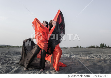 Two girls in black and red flowing fabric pose on sand dune 89437018