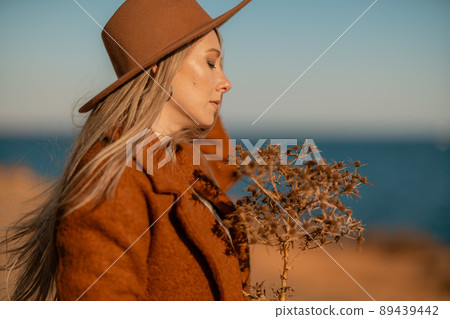 A woman walking along the coast near the sea. An elegant lady in a brown coat and a hat with fashionable makeup walks on the seashore 89439442