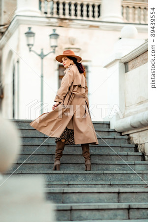 Outdoor fashion portrait of young elegant fashionable brunette woman, model in stylish hat, choker and light raincoat posing at sunset in European city. Outdoor fashion portrait of young elegant fashionable brunette woman, model in stylish hat, choker and light raincoat posing at sunset in European city. 89439554