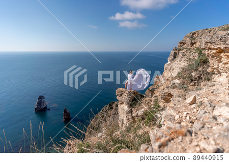 A beautiful young woman in a white light dress with long legs stands on the edge of a cliff above the sea waving a white long dress, against the background of the blue sky and the sea. 89440915