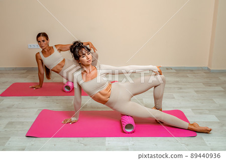 Fit athletic women in beige sportswear ride on a pink foam myofascial massage roller, massaging their muscles on the floor of a yoga mat in the gym. Active girls do a fitness workout in the gym. MFR 89440936