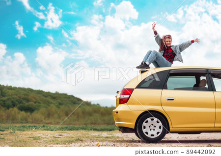 A happy teen girl is sitting on the roof of a yellow car, joyfully raising her hands up. Mountains and sky are in the background. Copy space. The concept of freedom and obtaining a driver's license 89442092