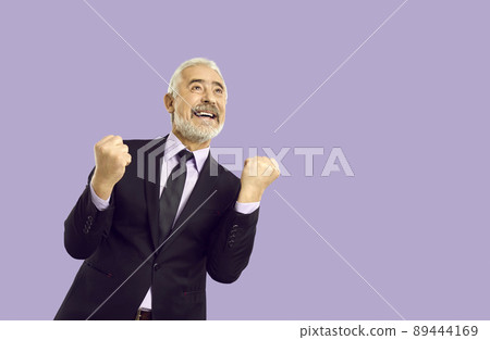 Happy emotional senior man in suit sincerely rejoices in his success isolated on purple background. 89444169