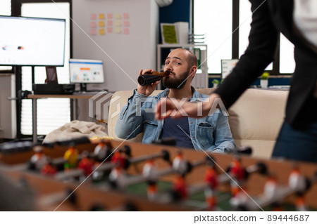 Caucasian man holding bottle of beer and waching foosball game, enjoying after work drinks with friends. Young person celebrating with alcohol, snacks, pizza, meeting with coworkers. 89444687