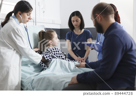 Pediatrician consulting sick little girl using stethoscope while worried parents waiting besides her. General practitioner examining ill daughter in pediatric clinic patient ward room. Pediatrician consulting sick little girl using stethoscope while worried parents waiting besides her. General practitioner examining ill daughter in pediatric clinic patient ward room. 89444728