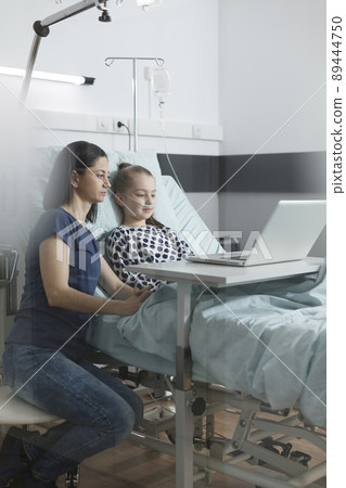 Young parent and unwell little daughter surfing web pages on laptop. Relaxed mother and daughter sitting in pediatric clinic patient ward room while using modern computer. 89444750