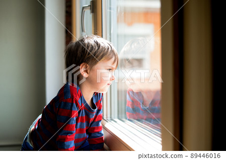 Kid boy sitting near window and looking outside. Serious child attentively looks out of the window 89446016