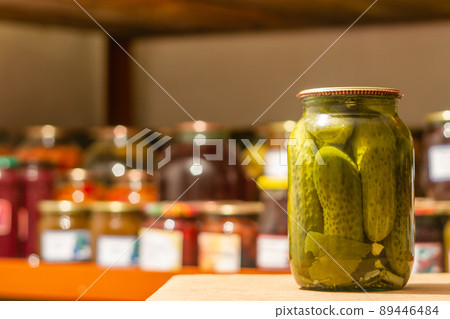 Homemade glass jar with pickled cucumbers in the cellar on the background of shelves with canned vegetables and fruits. Closeup, selective focus Homemade glass jar with pickled cucumbers in the cellar on the background of shelves with canned vegetables and fruits. Closeup, selective focus 89446484