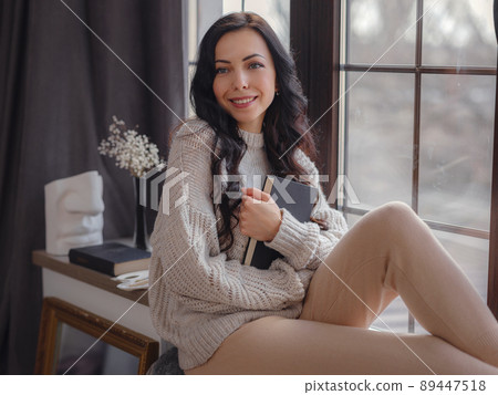 Beautiful young woman reading book near window at home 89447518