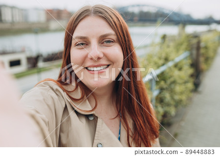 Happy cheerful young redhead woman in fashion dress posing outside. Woman taking selfie on urban background, summer time, traveling alone 89448883