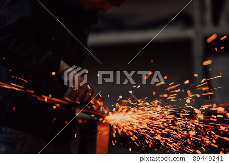 Heavy Industry Engineering Factory Interior with Industrial Worker Using Angle Grinder and Cutting a Metal Tube. Contractor in Safety Uniform and Hard Hat Manufacturing Metal Structures. Heavy Industry Engineering Factory Interior with Industrial Worker Using Angle Grinder and Cutting a Metal Tube. Contractor in Safety Uniform and Hard Hat Manufacturing Metal Structures. 89449241
