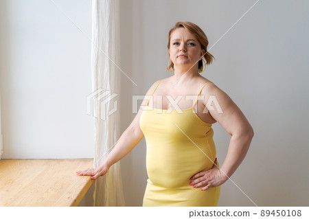 Adult plus size woman in tight-fitting yellow dress, makeup standing by window 89450108