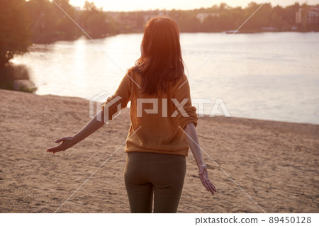 Happy woman relaxes with her arms out to sides on shore lake at sunset in rays Happy woman relaxes with her arms out to sides on shore lake at sunset in rays 89450128