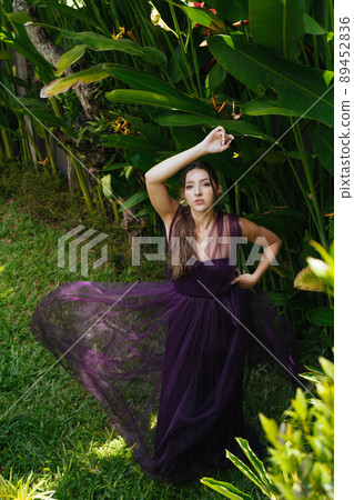 A girl in an evening long dress stands by a banana thicket posing for a photographer A girl in an evening long dress stands by a banana thicket posing for a photographer 89452836