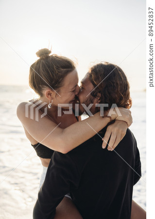 The man raised his beloved in his arms while she wrapped her arms around his neck laughing and kissing him against the background of the ocean and sand The man raised his beloved in his arms while she wrapped her arms around his neck laughing and kissing him against the background of the ocean and sand 89452837