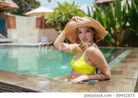A girl in a sun hat and swimsuit in the water pool leaned her elbow on the edge of the parapet and poses for a photo against a backdrop of tropical plants 89452873