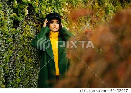 Beautiful girl in a stylish coat straightens on the head cap against the background of the wall with ivy 89452917