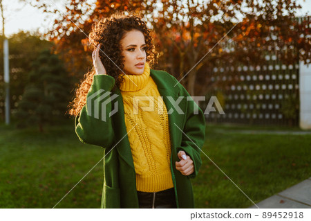 Luxury beauty in a bright yellow sweater and dark green coat stands on the green grass of the park Luxury beauty in a bright yellow sweater and dark green coat stands on the green grass of the park 89452918