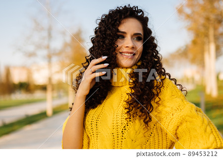 In a bright yellow sweater a beautiful woman with curly hair with a broad smile straightens her dark hair against the backdrop of autumn park 89453212