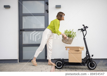 Woman carrying basket with herbs on a porch of her house with electric scooter and parcels nearby Woman carrying basket with herbs on a porch of her house with electric scooter and parcels nearby 89454547