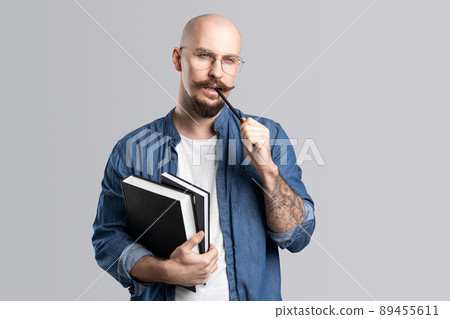Portrait of a handsome nerd guy on a gray background, holds a books and pen in his hand with a serious face and thinks. 89455611