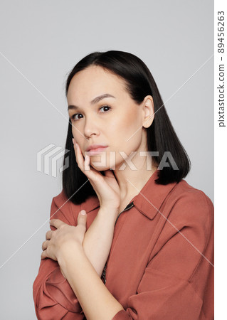 Portrait of serious young Asian woman with natural makeup touching own face against white background 89456263