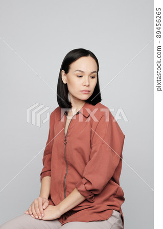 Sad young Asian woman in dark beige shirt sitting on stool and looking down Sad young Asian woman in dark beige shirt sitting on stool and looking down 89456265