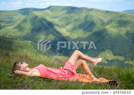 Young woman in red dress lying down on green grassy meadow on a warm sunny day in summer mountains enjoying view of nature. Young woman in red dress lying down on green grassy meadow on a warm sunny day in summer mountains enjoying view of nature. 89456834