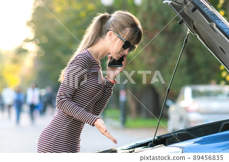 Young upset woman driver talking on mobile phone near a broken car with open hood waiting for help having trouble with her vehicle on a city street. Young upset woman driver talking on mobile phone near a broken car with open hood waiting for help having trouble with her vehicle on a city street. 89456835