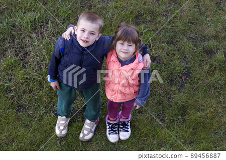 View from above of two cute children in casual clothes boy and girl standing on green grass, hugging each other over shoulders, looking up and shyly smiling. Happy childhood and friendship concept. View from above of two cute children in casual clothes boy and girl standing on green grass, hugging each other over shoulders, looking up and shyly smiling. Happy childhood and friendship concept. 89456887